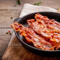 Hot fried crispy bacon slices in skillet on wooden background.